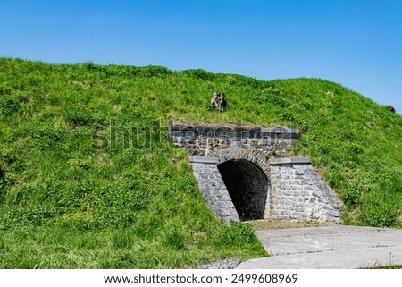 Underground tunnel leading to one of complexes in Charlemont fortress, green grass on hill against blue sky, thick stone walls and arched entrance, sunny day in Givet, Ardennes department, France