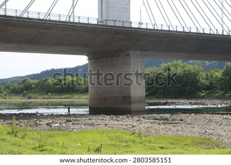 The underside of a large bridge and its central concrete support dominate the frame above a low-water riverbed. A small distant person is visible near the water, with rocks, trees, and green banks completing the outdoor infrastructure scene.