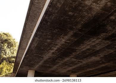 The underside of a modern elevated highway structure or concrete bridge, dark, textured surface, possibly due to aging or moisture, the structure is supported by concrete pillars