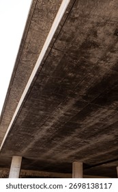 The underside of a modern elevated highway structure or concrete bridge, dark, textured surface, possibly due to aging or moisture, the structure is supported by concrete pillars