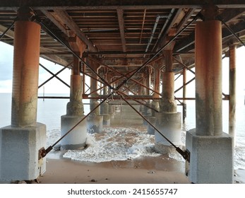 Underside of a pier with repeating patterns of columns and beams disappearing into the distance and waves underneath