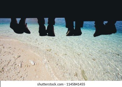 Underside view of dangling Four people legs with wearing trekking shoes sitting on the edge of wooden board-walk against a background of Blue sea.