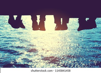 Underside view of dangling Four people legs with wearing trekking shoes sitting on the edge of wooden board-walk against a background of Blue sea.