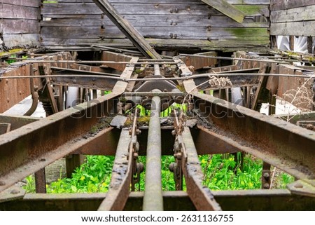 Underside view of a rusted train car frame and suspension components in an old, decaying railway wagon. Corroded beams, mechanical linkages, and overgrown vegetation create a sense of industrial aband