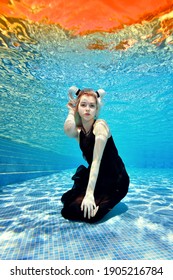 Underwater Portrait Of A Blonde Girl Who Sits And Poses For The Camera Underwater At The Bottom Of An Outdoor Pool In A Black Dress With White Horns On Her Head On A Sunny Day. Fashion Photography.