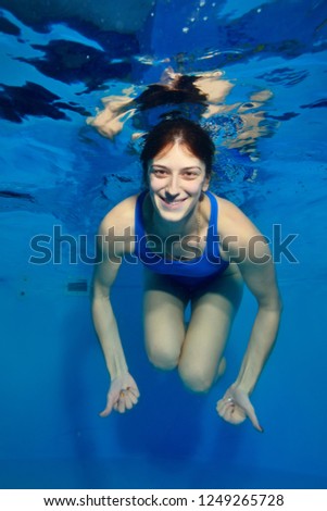 Underwater portrait of a girl-coach, who swims under the water in the pool on a blue background, looking at the camera and smiling. Conceptual realism. Vertical orientation