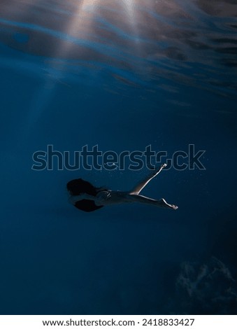Underwater shoot of beautiful sportive girl in bodysuit swimming in water through sunbeams. Fantasy mermaid against water surface background with rays of lights.