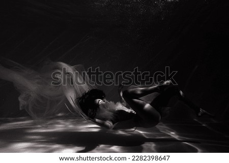 Underwater shoot of beautiful woman in white flying transparent dress relaxing in water in sunbeams. Fantasy mermaid against water surface background with rays of lights. Black and white image.