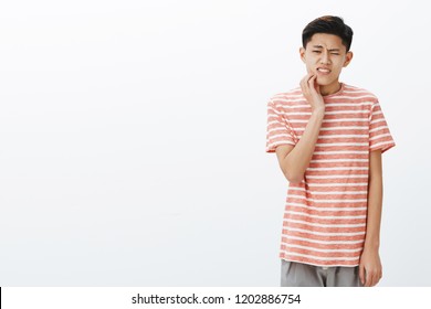 Uneasy Attractive Young Asian Male Student Having Tooth Decay Touching Cheek Reacting On Pain, Having Rotten Teeth, Suffering Painful Feeling While Waiting In Queue To Dentist Over Grey Wall