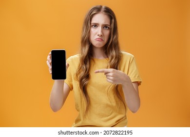 Uneasy and moody cute young woman with natural long wavy hair showing smartphone pointing at cellphone screen displeased and glooy being upset gadget do not work, posing over orange wall