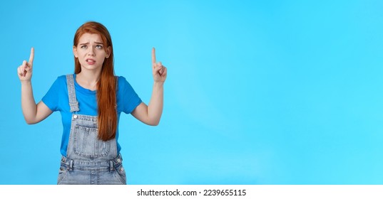 Uneasy worried cute redhead female coworker feel unsure insecure, grimacing displeased intense worried expression, look pointing up, top copy space, have hesitations, stand blue background. - Shutterstock ID 2239655115