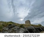 Unedited photo of a historic stone tower on a rocky hilltop in Tuscany, Italy, under a dramatic, cloudy sky. The image captures the ancient and rugged beauty of the region’s architectural heritage