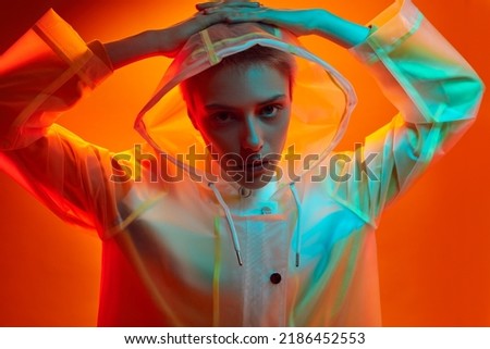 Unemotional young female model with short blond hair in trendy transparent hooded raincoat, touching head and looking at camera against orange background in studio with blue neon light Stock photo © 