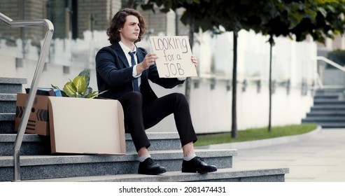 Unemployed businessman in despair sits on stairs outdoors next to a box with personal belongings, holding cardboard sign banner that says looking for a job. Fired office worker seeks help.