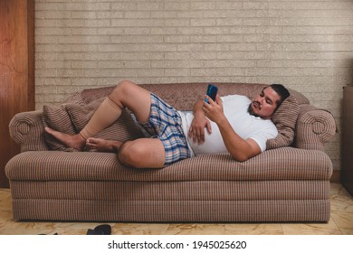 An unemployed and lazy man watching videos, playing mobile games or on social media on his cellphone all day while lying on the couch recovering from a calf injury.