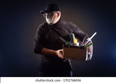 An Unemployed Man With A Mask On His Face And A Box Of Office Supplies. Unemployment. Job Loss Due To The Coronavirus Epidemic. Ruin. Bankruptcy. Depression From Job Loss.