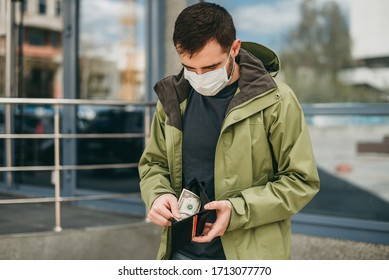 Unemployed Young Man In A Medical Mask Holding A Wallet With A Few Dollars Outside, No Money, Crisis, Poverty, Hardship. Quarantine, Coronavirus, Isolation.