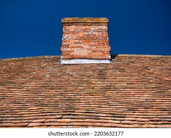 Uneven, weathered, old clay tiles and chimney on a traditional cottage roof in England, UK, taken on a sunny day with a blue sky. Lead flashing is visible at the joint of the chimney and roof.  - Shutterstock ID 2203632177