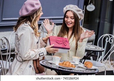 Unexpected Surprise From Best Friend. Side View Of Beautiful French Woman Giving Present To Her Surprised Friend While Sitting In French Cafe. Celebrating Birthday Together. Birthday Gift Box.