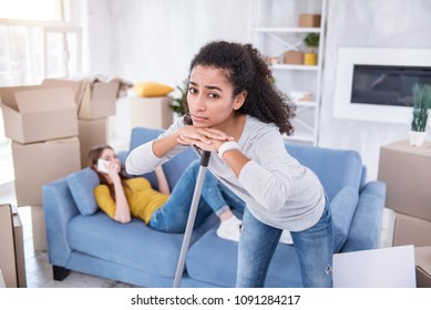 Unfair Life. Pretty Curly Girl Cleaning The Floor In The New Apartment While Her Roommate Lying On The Sofa And Doing Nothing But Talking On The Phone