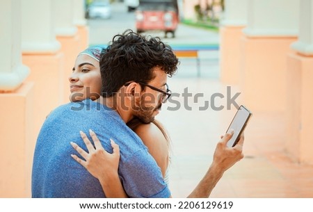 Unfaithful man looking at the cell phone while hugging his girlfriend outdoors. Unfaithful boyfriend hugging his girlfriend and looking at the cell phone. Concept of unfaithful man using cell phone