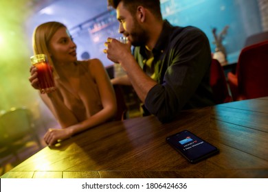Unfaithful man spending time with another woman in the bar, enjoying drinks and conversation. Cheating, infidelity concept. Focus on smartphone, wife or girlfriend is texting. Horizontal shot