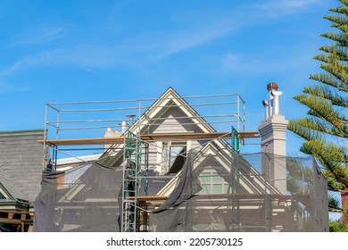 Unfinished House Exterior With Scaffolding Covered With Net In San Francisco, California. View Of A Peak Of An Under Construction House With Chimney Near The Pine Tree On The Right.