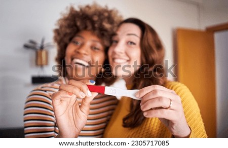 Unfocused smiling young lesbian multiracial couple looking at camera and holding positive pregnancy test. Focus hands predictor. People in love expecting a baby. Future happy homosexual LGBT family. 
