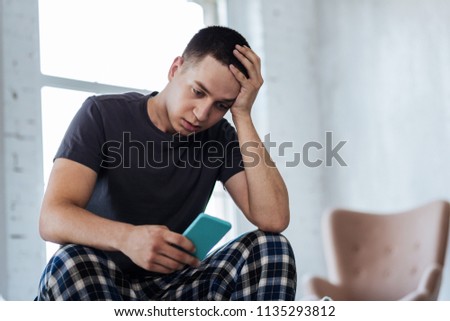 Unhappiness. Upset young man sitting at home and holding his phone