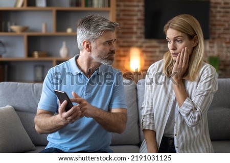 Unhappy adult caucasian man with beard shows phone to shocked lady, woman suffers from gadget addiction in room interior. Distrust, problems in relationships, cheating and gambling, waste of money