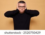 Unhappy adult male wearing black jumper and spectacles grimacing in irritation from noisy environment feeling troubled looking at camera with sad expression and pout lips isolated over beige 