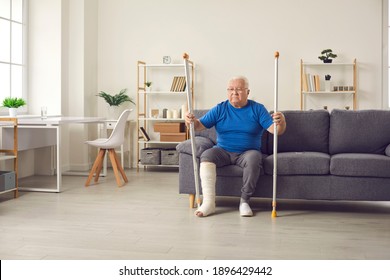 Unhappy Adult Patient With Physical Injury Sitting On Sofa At Home And Holding Crutches. Sad Thoughtful Senior Man With Broken Leg In Cast Wants To Stand Up From Couch And Walk In The Living-room