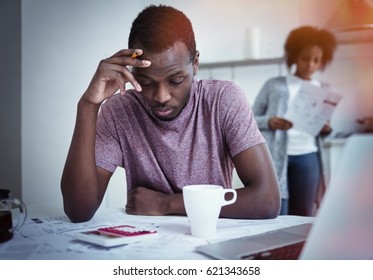 Unhappy african american family financial stress: black man sitting at kitchen, reading notification informing that bank denied to prolong loan term, his wife standing with bills in background