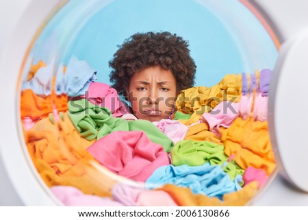 Unhappy African American woman with curly hair has tired expression does laundry at home drowned in multicolored clothes shows only head feels discontent. Sulking dissatisfied female model at washer