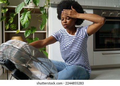 Unhappy African woman touching forehead having hot flashes using electric fan to cool off, overheated black female sitting in front of air cooler at home, surviving heatwave without air conditioning
