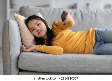 Unhappy Asian Kid Pretty Long-haired School-aged Girl Laying On Couch At Home, Holding Remote Controller, Watching TV Alone, Searching Nice Movie Or Cartoon. Kids Leisure, Domestic Entertainment