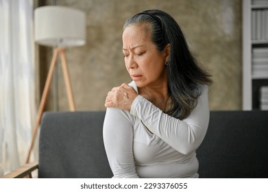 Unhappy Asian mature woman suffering from shoulder pain, massaging her shoulder while resting on sofa in her living room. - Powered by Shutterstock