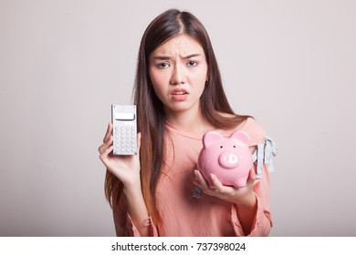 Unhappy Asian woman with calculator and piggy bank on gray background