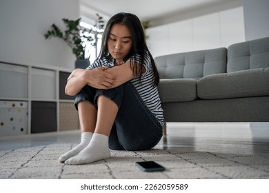 Unhappy Asian woman think about troubles looks on mobile phone wait call sadly sits on floor at home. Upset young female suffering from breakup. Anxious Korean girl blogger scared bullying by haters. - Powered by Shutterstock