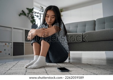 Unhappy Asian woman think about troubles looks on mobile phone wait call sadly sits on floor at home. Upset young female suffering from breakup. Anxious Korean girl blogger scared bullying by haters.