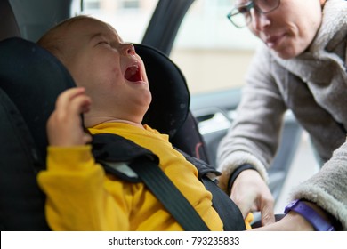 Unhappy Baby Boy Crying In Car Seat While His Mother Is Trying To Fasten The Safety Belts