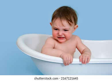 Unhappy Baby Toddler Boy Is Sitting In A White Tub On A Studio Blue Background. Crying Child At The Age Of One Year