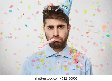 Unhappy birthday guy with stubble feeling sad and disappointed because nobody came to celebrate his anniversary, blowing party horn all alone, confetti flying around him. People and celebration