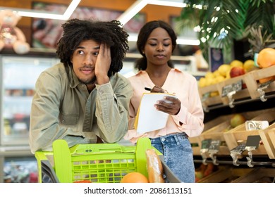 Unhappy Black Family Couple Counting Food Prices Struggling From Financial Crisis Calculating Expenses And Taking Notes In Grocery Shopping List Buying Products In Supermarket Indoor