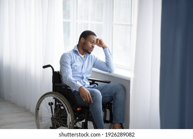 Unhappy black handicapped guy looking out window, feeling sad and desperate at home, copy space. African American man in wheelchair suffering from disability depression, indoors