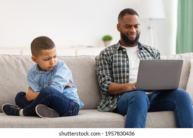 Unhappy Black Kid Boy Sitting Near Reluctant Indifferent Father While He Using Laptop Not Paying Attention To Offended Little Son Sitting On Sofa At Home. Family Problems, Bad Relationship With Dad