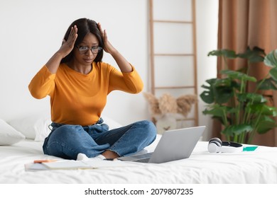 Unhappy Black Woman Looking At Laptop Touching Head In Shock Having Problem With Computer Or Bad Internet Connection Sitting In Bedroom At Home. Freelance Issues Concept