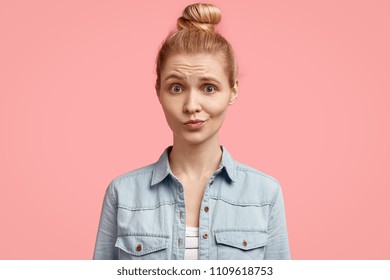 Unhappy blonde European female with discontent expression, frowns face, recieves bad news from interlocutor, stands against pink background. Disappointed young woman in denim jacket has no make up