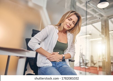 Unhappy Blonde Lady Feeling Pain In Her Stomach While Sitting In Chair At Work