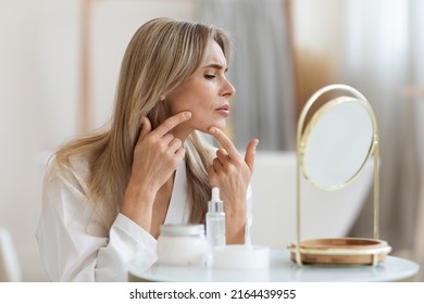 Unhappy Blonde Middle Aged Lady In White Silky Bathrobe Sitting In Front Of Mirror In Bathroom, Removing Blackheads From Her Chin Skin, Copy Space. Acne, Pimples, Problem Skin Care Concept